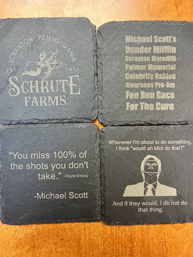 Four slate coasters with engraved text and branding on a wooden surface. the office inspired gift